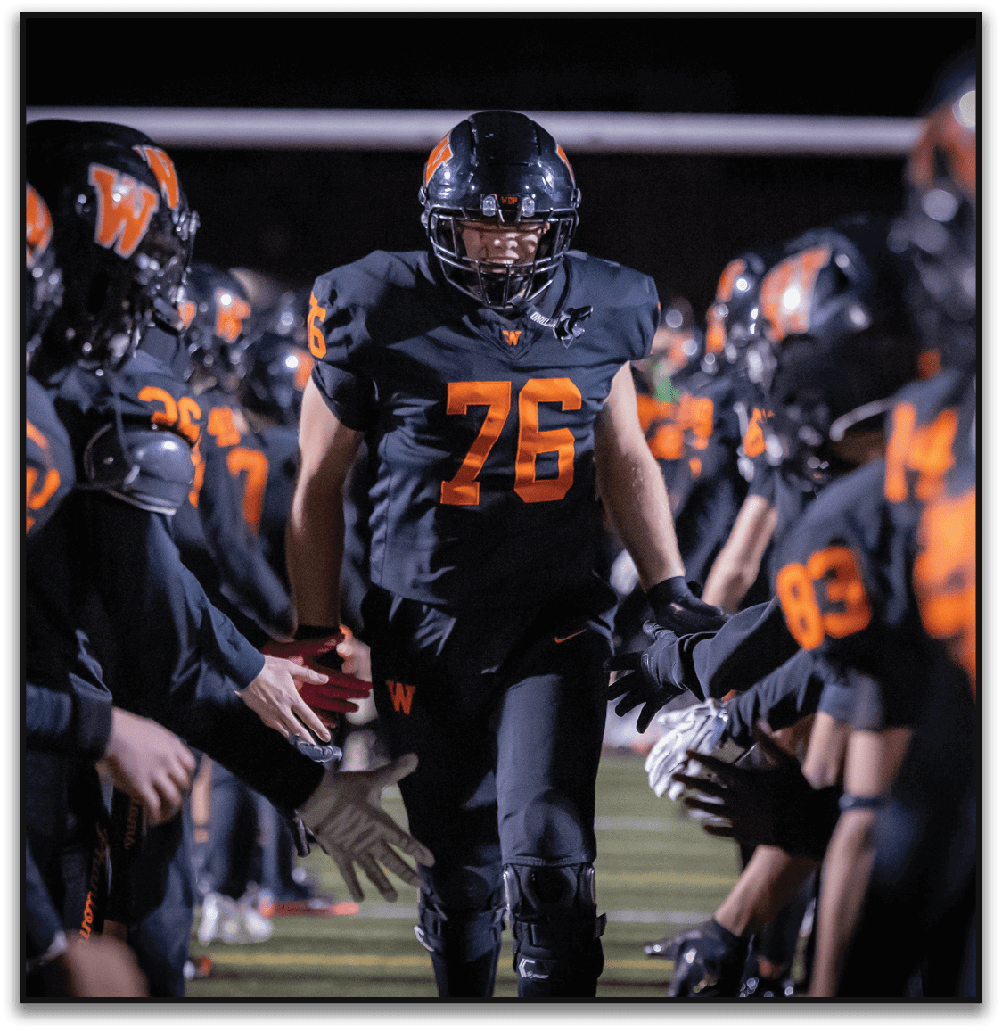 A person wearing a black football uniform with the number 76 is walking between two lines of teammates who are reaching out to give high fives on a football field at night. Description generated by AI