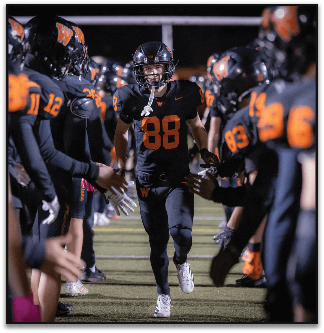 A person wearing a black football uniform with the number 88 is running through a tunnel formed by teammates in similar uniforms on a football field at night. Description generated by AI