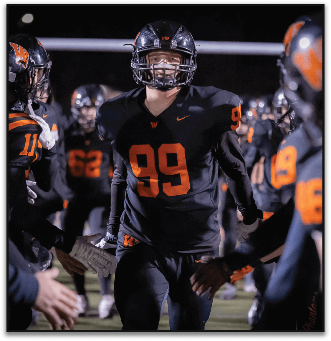 A person wearing a black football uniform with the number 99 is running through a line of teammates who are reaching out to give high fives on a lit football field at night. Description generated by AI