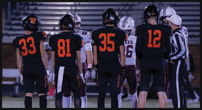 Five people in football uniforms stand on a field facing away from the camera with a referee and two other players in the background. Description generated by AI
