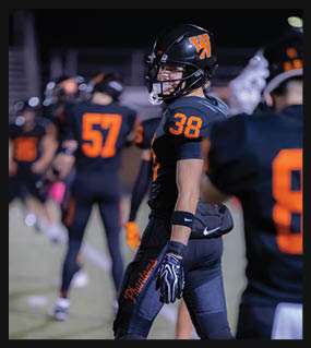 A person wearing a black football uniform with the number 38 in orange and a helmet is standing on a field with other players in the background. Description generated by AI