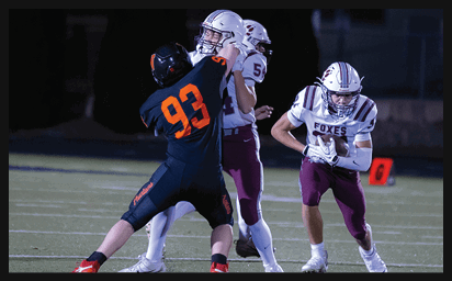 Three people in football uniforms are on a field with one player in a dark uniform attempting to block or tackle two players in white and maroon uniforms, one of whom is holding a football. Description generated by AI