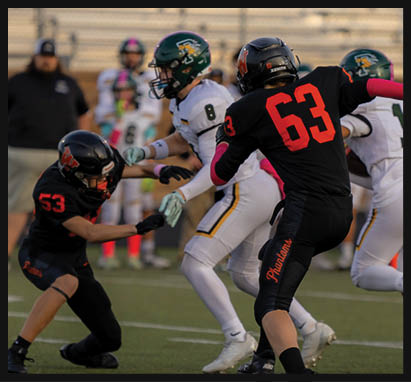 Two people in black football uniforms with red numbers are on a field, one standing and the other crouching, while others in white uniforms and a person in casual clothing stand in the background. Description generated by AI