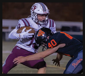 Two people in football uniforms are engaged in a tackle during a game, with one holding the ball tightly while the other reaches to stop them. Description generated by AI