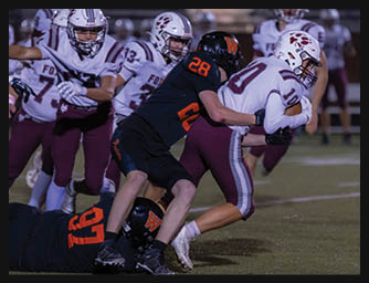 Two people in football uniforms are engaged in a tackle on a grassy field during a game, with other players visible in the background. Description generated by AI