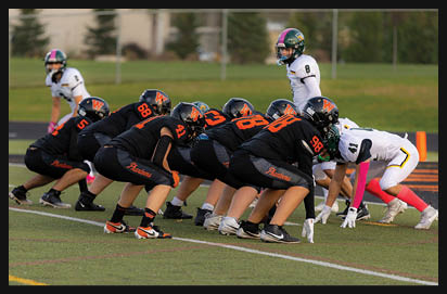 A group of athletes in black and orange uniforms are lined up in a football stance facing a smaller group of athletes in white uniforms on a green field. Description generated by AI