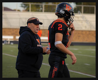 A person wearing a black hoodie and visor is standing next to a person in a black and orange football uniform with a helmet on a football field. Description generated by AI