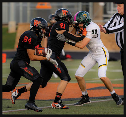A person in a black football uniform with the number 2 is holding a football and preparing to throw while another person in a white football uniform with the number 8 approaches on a field with a blurred person in an orange vest in the background. Description generated by AI