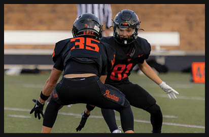 Two people in black football uniforms with orange numbers are positioned facing each other on a field, appearing ready for a play. Description generated by AI