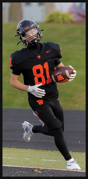 A person wearing a black football uniform with the number 81 is running on a field while holding a football. Description generated by AI