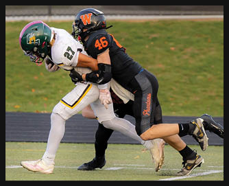 Two people in football uniforms are engaged in a tackle on a grassy field with one person in white running while the other in black holds them from behind. Description generated by AI