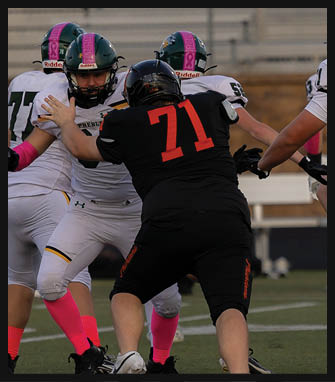 Two people in football uniforms are engaged in a blocking action on a field, with one wearing a black jersey numbered 71 and the other in a white jersey with green and pink accents. Description generated by AI