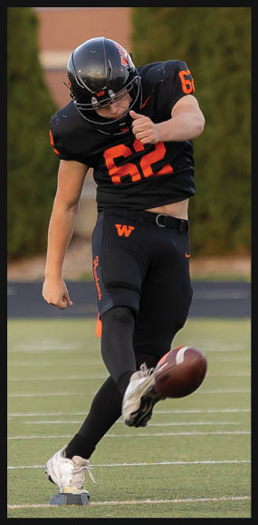 A person in a black and orange football uniform is kicking a football on a field. Description generated by AI