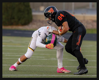 Two people in football uniforms are engaged in a tackle on a field, with one person in black and orange gear holding the other person in white and green gear who is bent forward. Description generated by AI