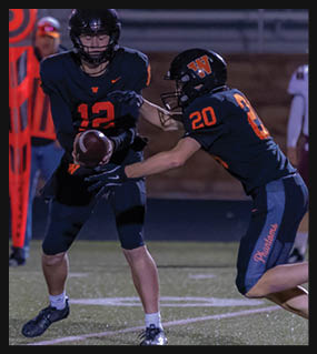 Two people in football uniforms are engaged in a play on a field, with one person holding the ball and the other reaching out to block or tackle. Description generated by AI