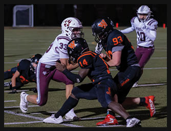 Two people in black football uniforms tackle a person in a white and maroon football uniform on a field while another person in white and maroon stands in the background. Description generated by AI