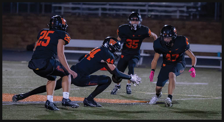 Four people in black and orange football uniforms are on a field at night with one person running while the others prepare to catch or block. Description generated by AI