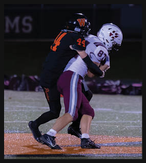 Two people in football uniforms are engaged in a tackle on a field with artificial turf under stadium lighting. Description generated by AI