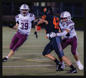 Two people in football uniforms are engaged in a play on a field, with one person wearing a dark uniform holding the ball and another person in a white and maroon uniform approaching. Description generated by AI