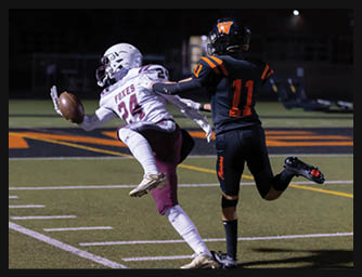 Two people in football uniforms are competing on a field with one person in white and maroon reaching out to catch a football while the other in black and orange attempts to defend. Description generated by AI