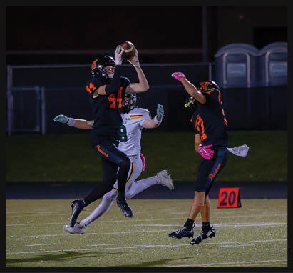 Three people in football uniforms are on a field at night with one person jumping to catch a football while the others reach toward the ball. Description generated by AI