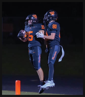 Two people in black football uniforms with orange numbers and helmets celebrate on a field at night, with one holding a football. Description generated by AI