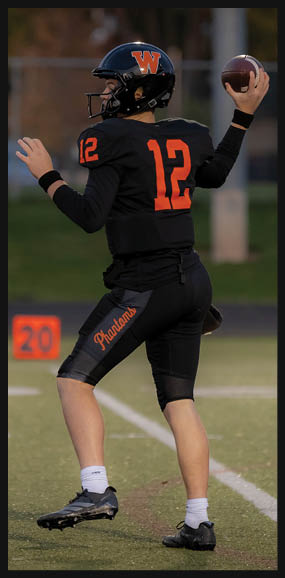 A person wearing a black football uniform with the number 12 and a helmet is preparing to throw a football on a field near a marker showing the number 20. Description generated by AI