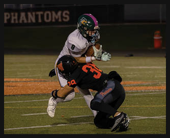 A person in a white football uniform with a green helmet is running with the ball while being tackled by another person in a black uniform with an orange number 33 on a football field at night. Description generated by AI