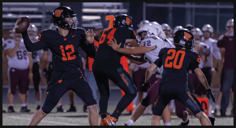 A group of people in football uniforms are playing on a field with one person in a black uniform preparing to throw a football while others block or defend. Description generated by AI