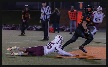 A person in black and orange football gear is running with the ball while another person in white and maroon football gear is diving on the ground attempting to tackle them on a football field. Description generated by AI