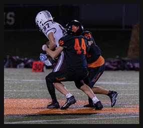 Two people in football uniforms are engaged in a tackle during a nighttime game on a field. Description generated by AI