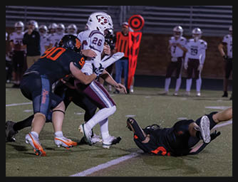 Two people in dark uniforms tackle a person in a white and maroon football uniform on a field during a nighttime game. Description generated by AI