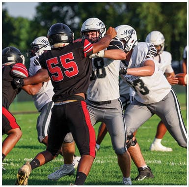 People wearing football uniforms are engaged in a play on a grassy field with one person holding a football and others blocking or tackling. Descripci n generada con IA