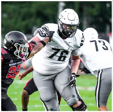 Two people in football uniforms are engaged in a play on a grassy field, with one person in black and red attempting to block or tackle the other in white and gray. Descripci n generada con IA