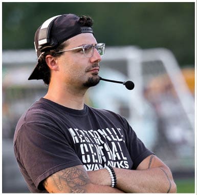 A person wearing a backward cap, glasses, and a headset stands with arms crossed on a sports field near a goalpost, watching intently. Descripci n generada con IA