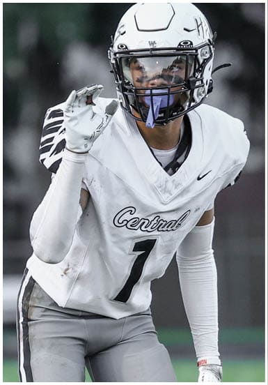 A person wearing a white football uniform with the number one and a helmet is standing on a grassy field making a hand gesture. Descripci n generada con IA