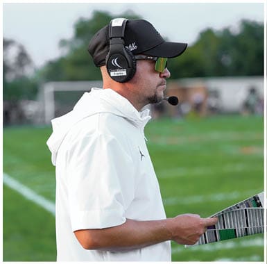 Person wearing a white hooded shirt and headset is holding a folded paper while standing on a grassy sports field. Descripci n generada con IA