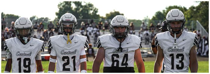 Four people wearing white football uniforms with helmets stand side by side on a grassy field with a crowd in the background. Descripci n generada con IA