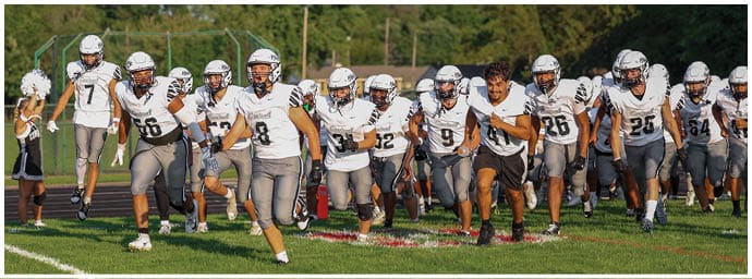 A group of people wearing football uniforms and helmets are running energetically on a grassy field during daylight. Descripci n generada con IA