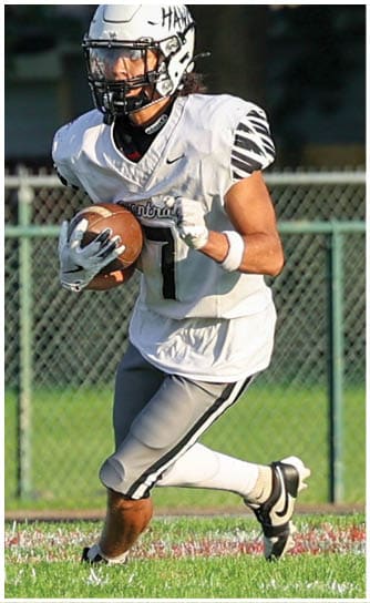 An adult wearing a white football uniform and helmet is running on a grassy field while holding a football. Descripci n generada con IA