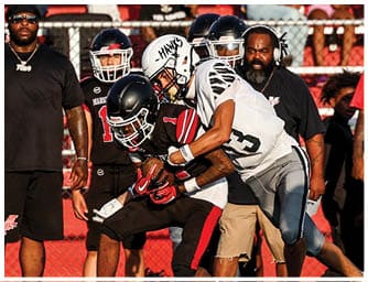 Two people in football uniforms are engaged in a play on a grassy field with one person holding the ball while the other attempts to tackle. Descripci n generada con IA
