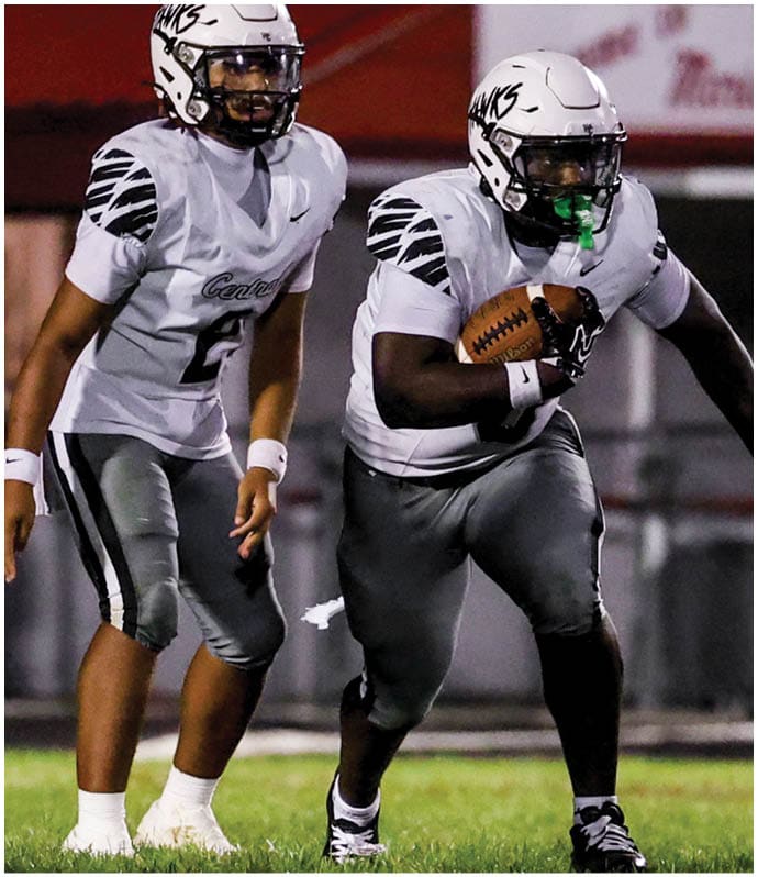 Two people in football uniforms and helmets are on a grassy field at night, with one holding a football and the other standing behind. Descripci n generada con IA