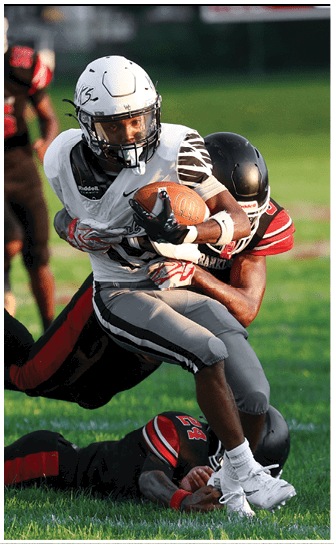 Two people in football uniforms are engaged in a tackle on a grassy field during a game. Descripci n generada con IA
