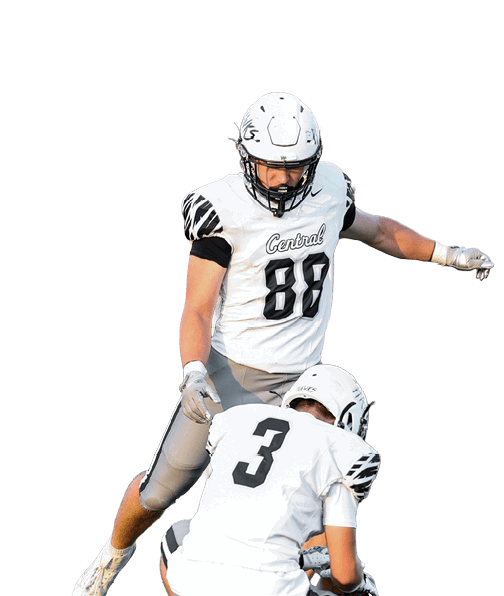 Two people in football uniforms are on a field with one holding the ball on the ground and the other preparing to kick it. Descripci n generada con IA
