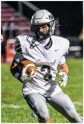 A person wearing a white football uniform with the word Central and the number 3 is running on a grassy field while holding a football. Descripci n generada con IA