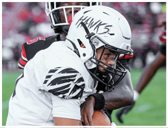 Two people in football uniforms are engaged in a close play on a grassy field with a blurred crowd in the background. Descripci n generada con IA