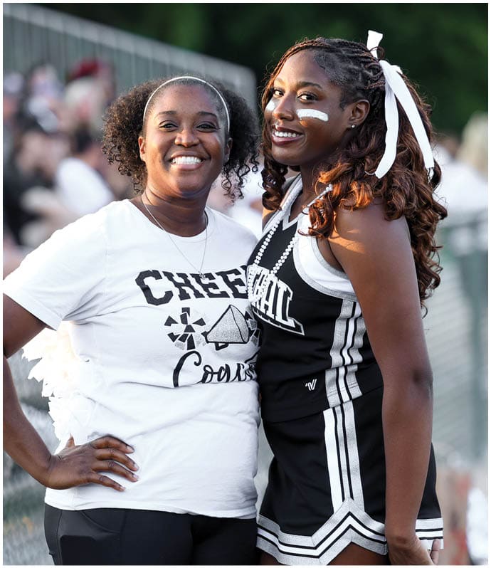 Two adults smiling and posing together at an outdoor event, one wearing a cheer coach t-shirt and the other in a cheerleading uniform with face paint and a ribbon in their hair. Descripci n generada con IA