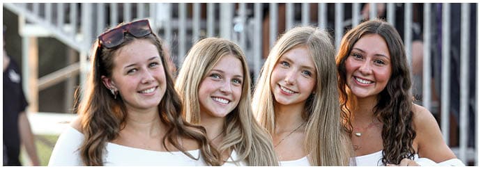 Four smiling adults with long hair stand closely together outdoors with a metal fence in the background. Descripci n generada con IA