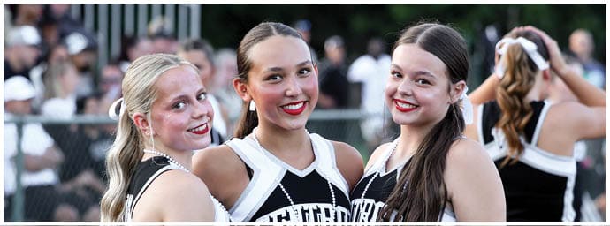 Three smiling adults wearing matching cheerleading uniforms stand close together outdoors with a crowd in the background. Descripci n generada con IA
