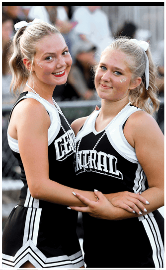Two adults wearing matching cheerleading uniforms and pearl necklaces smile while posing closely together. Descripci n generada con IA